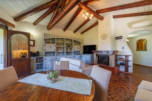 A cozy living room with a fireplace, wooden beams, and a dining area with a table set for two. - Villa Baja (Baja Sardinia)