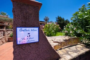 Un cartel en un pilar de piedra con el texto 'Conteggio, lettria 5 Via 3 Monti Baio Cerdeña'.