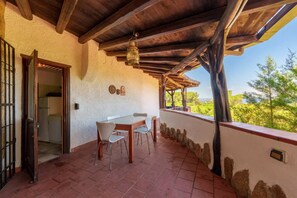 A covered patio with a table and chairs, a view of greenery, and a rustic architectural style. - Villa Baja (Baja Sardinia)