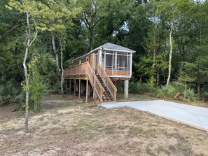 A wooden deck house with a staircase leading to a covered porch, surrounded by dense trees. - Bayou Bengal Bungalow Fire Pit-Grill-Screened Porch-Near LSU & Bars Restaurants (Baton Rouge)