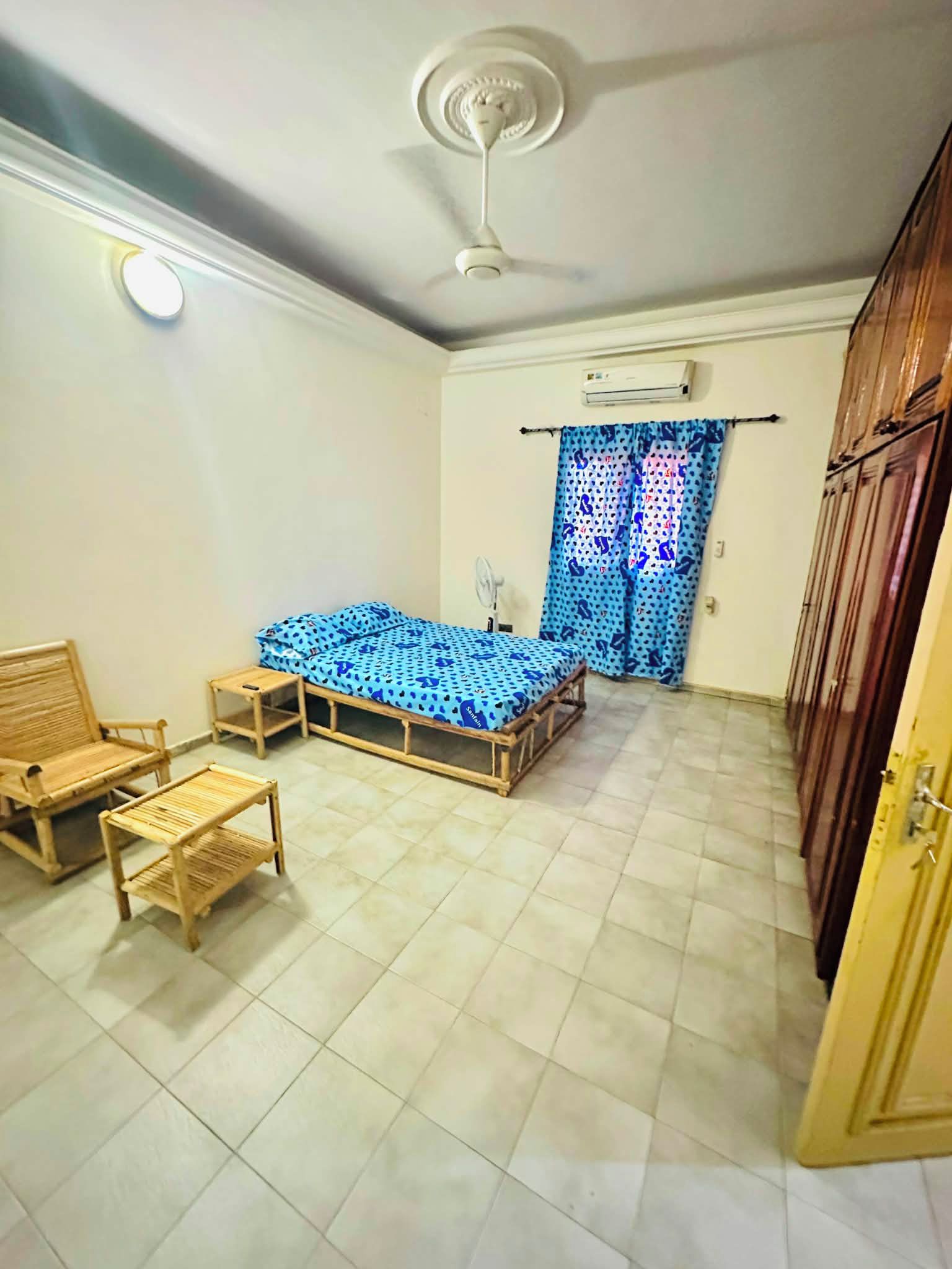 Een slaapkamer met een bed, twee houten stoelen, een tafeltje, een plafondventilator, een airconditioning en een kledingkast.