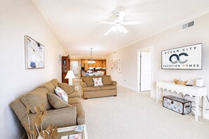 Ein geräumiger Wohnbereich mit zwei Sofas, einem Esstisch und einer Küche im Hintergrund. Ein Deckenventilator ist sichtbar, und ein Schild an der Wand wirbt für Ocean City Vacation Homes.