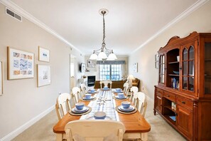 A dining room with a long wooden table set for a meal, flanked by white chairs, a wooden cabinet with glass doors, and framed artwork on the walls. - Laguna Vista 304 (Ocean City)