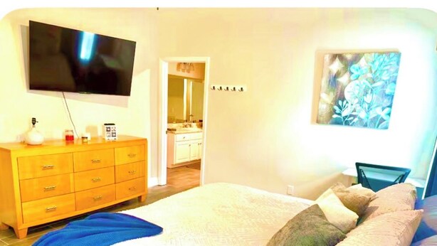 A bedroom with a bed, a TV mounted on the wall, a dresser, and a bathroom visible through an open door.