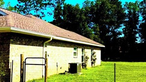 A single-story brick building with a covered porch, a fenced yard, and trees in the background.
