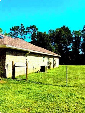 A single-story brick building with a covered porch, a fenced yard, and trees in the background.