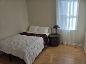 A bedroom with a bed, a nightstand, a lamp, and a window with sheer curtains.
