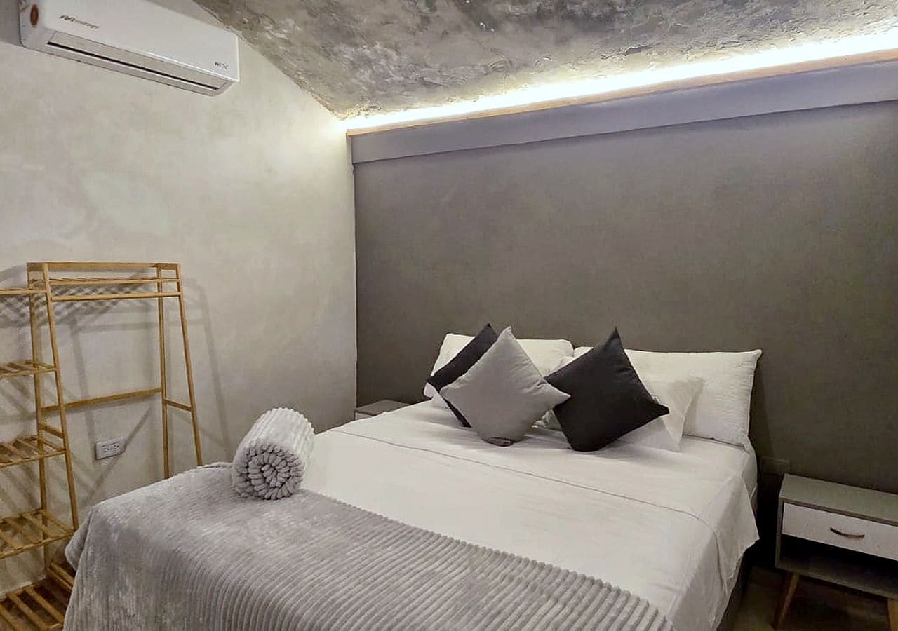 A hotel room with a bed, a bedside table, a wall-mounted air conditioner, and a wooden ladder against the wall.