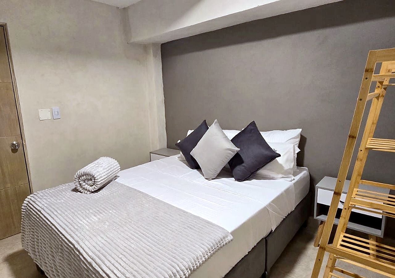 Una cama bien hecha con una colcha blanca y almohadas, una escalera de madera y una mesita de noche blanca con una lámpara.