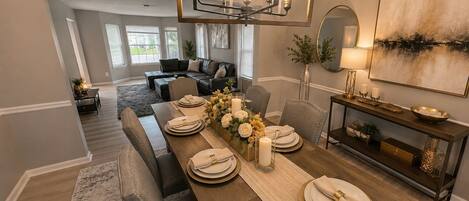 A dining area with a long wooden table set for six, a chandelier, and a large painting on the wall.