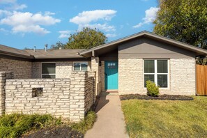 A two-story house with a stone facade, a blue front door, and a small front yard with grass and a bush. - 3BR Retreat in Austin — Massage chair & Firepit (Austin)