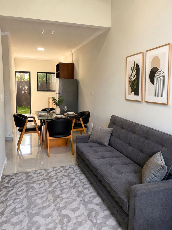 A modern living room with a grey sofa, a dining area with a table and chairs, and two framed artworks on the wall.