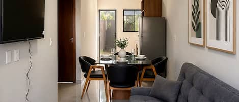 A modern living room with a grey sofa, a flat-screen TV mounted on the wall, and a dining area with a table and chairs.