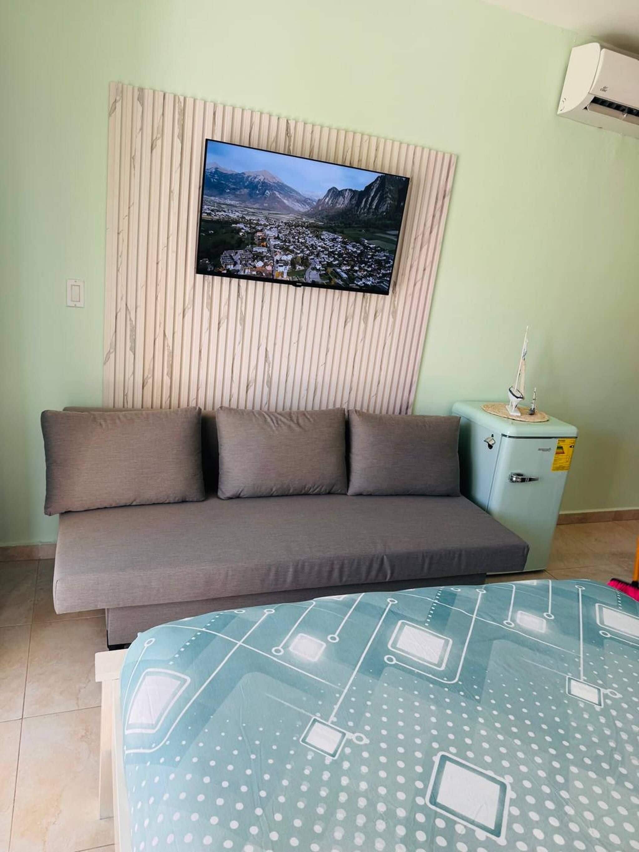 A room with a sofa, a television mounted on a wall, a small refrigerator, and a table with a patterned tablecloth.