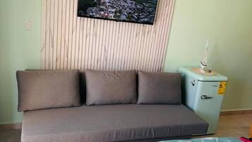 A room with a sofa, a television mounted on a wall, a small refrigerator, and a table with a patterned tablecloth.