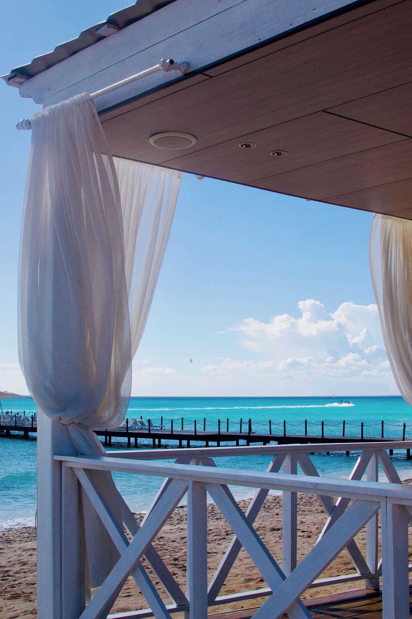 Vista a la playa con una terraza de madera y cortinas blancas.