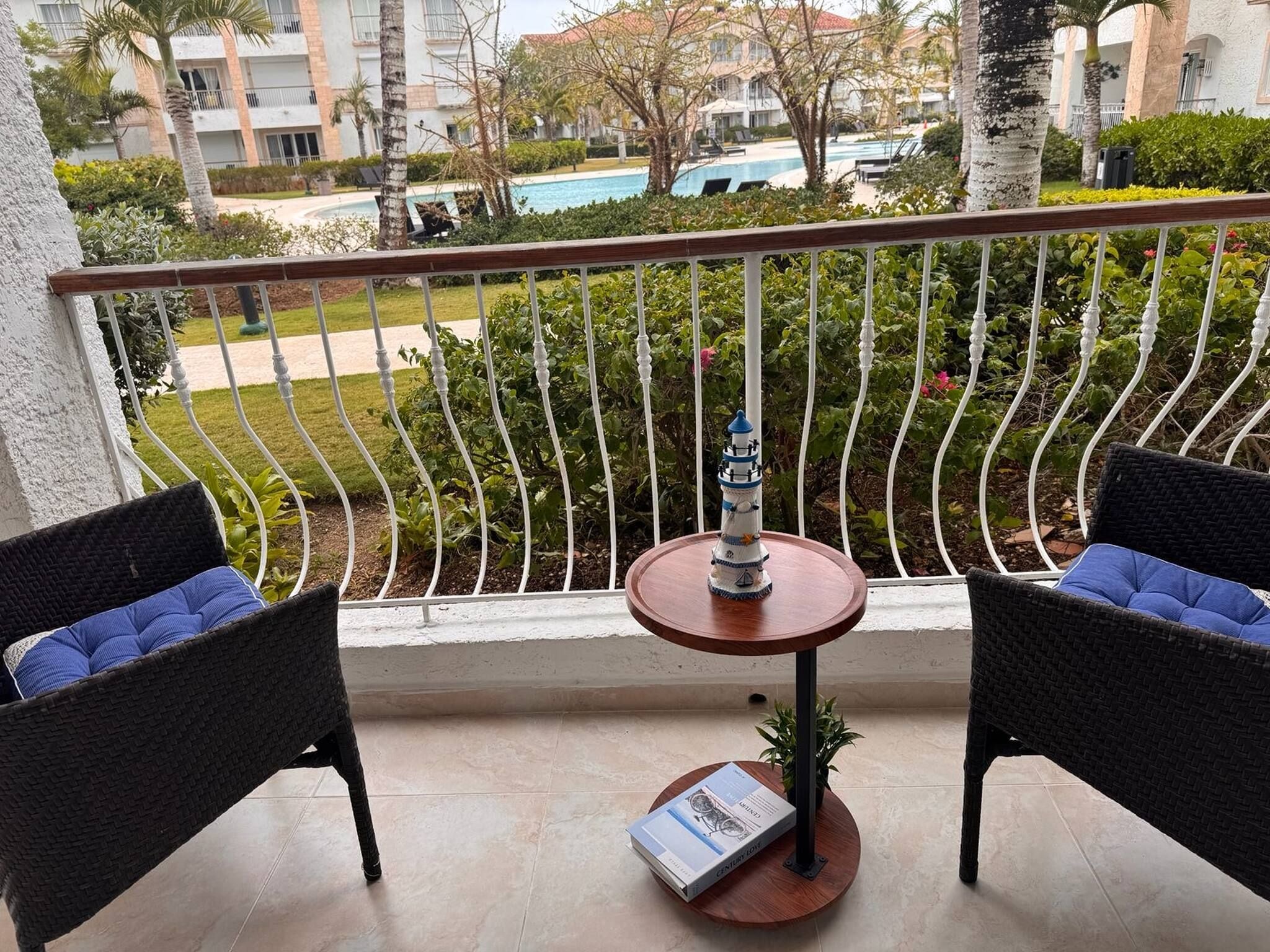 A balcony with two wicker chairs, a small table, and a book. A pool and buildings are visible in the background.