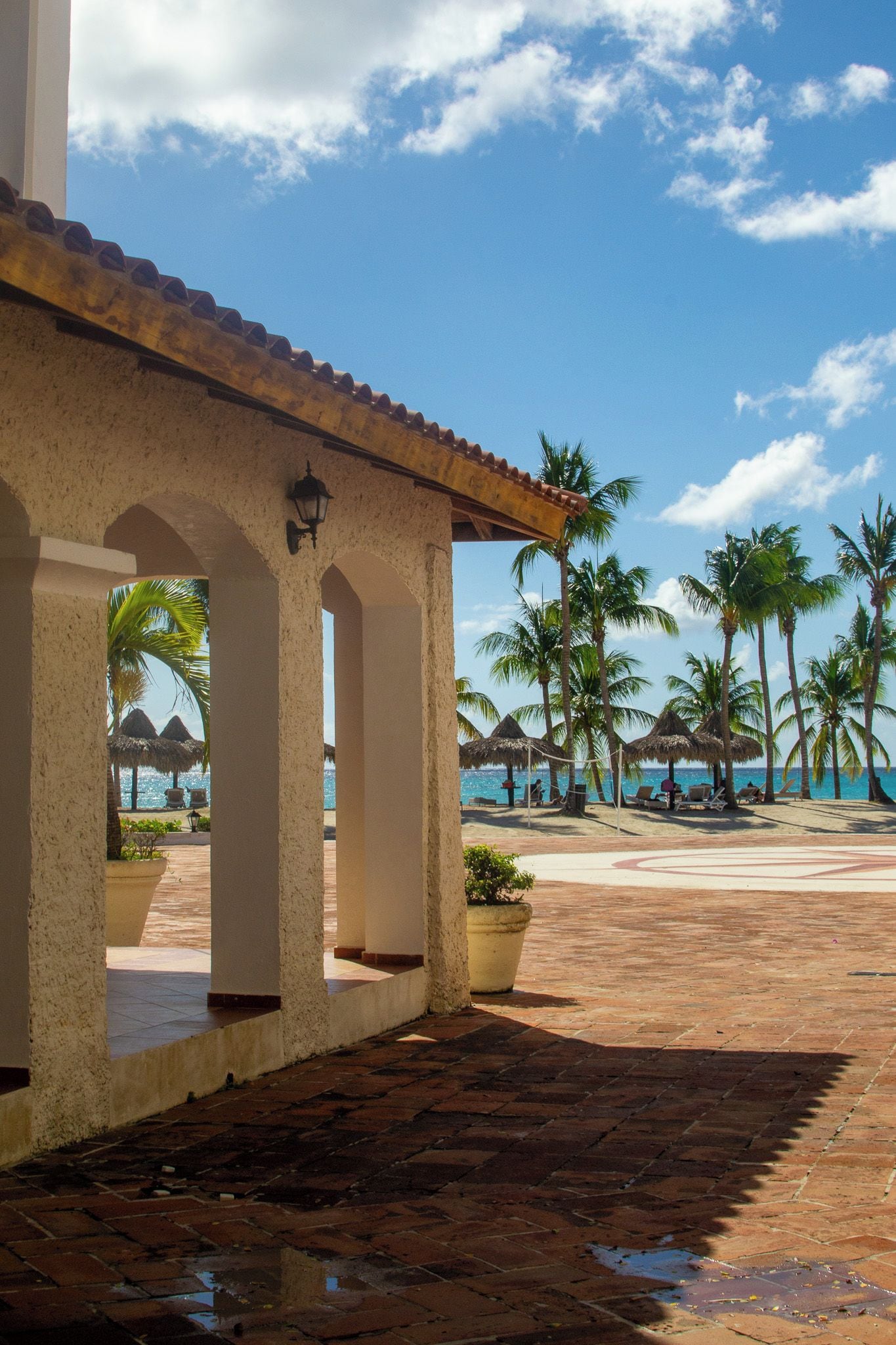 A covered outdoor area with a tiled floor, palm trees, and a view of the ocean.