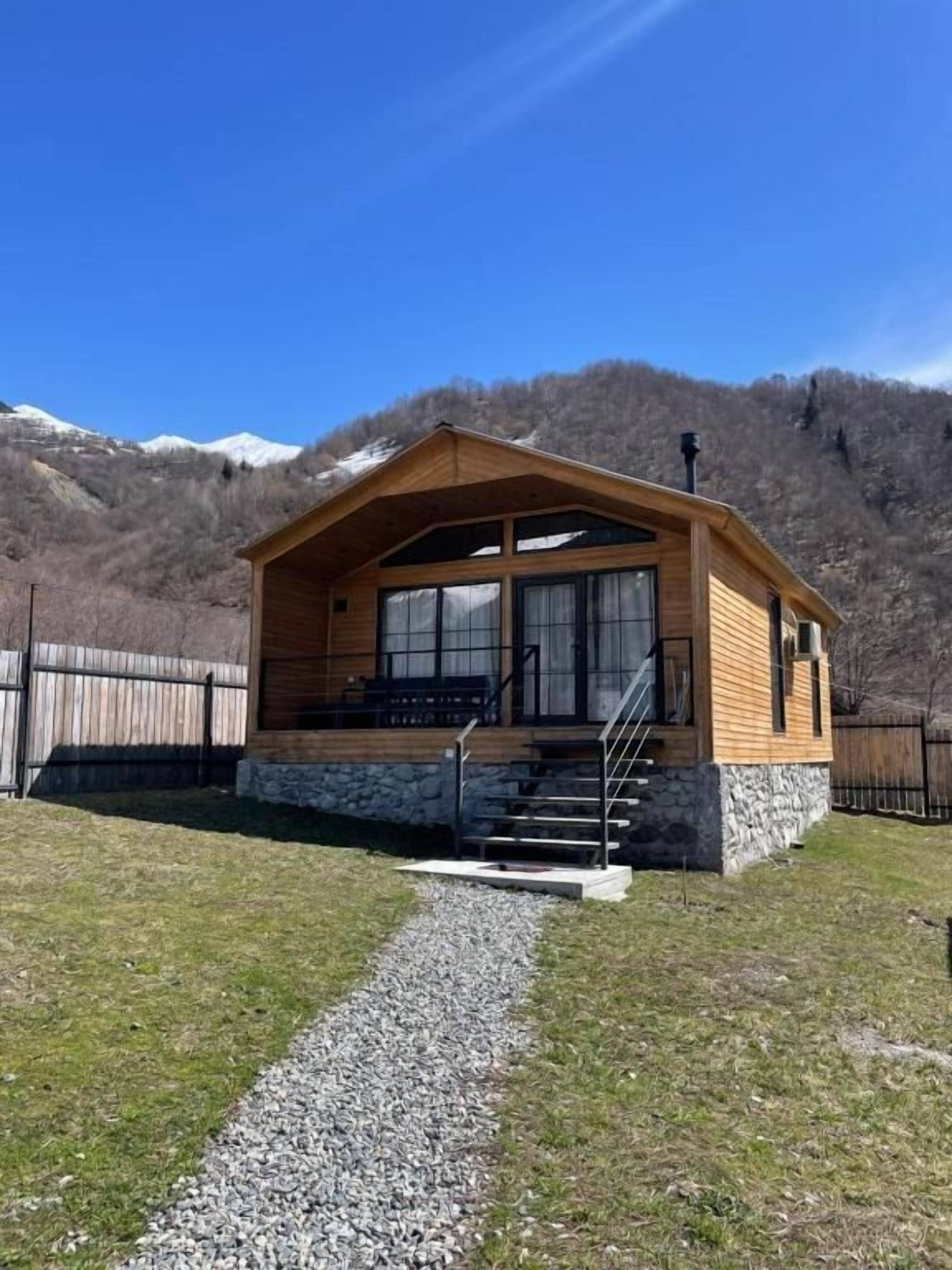 Une cabane en bois, construite sur des fondations en pierre, avec un escalier menant à l’entrée et offrant une vue dégagée sur les montagnes environnantes.