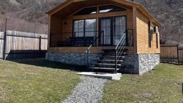 Une cabane en bois, construite sur des fondations en pierre, avec un escalier menant à l’entrée et offrant une vue dégagée sur les montagnes environnantes.