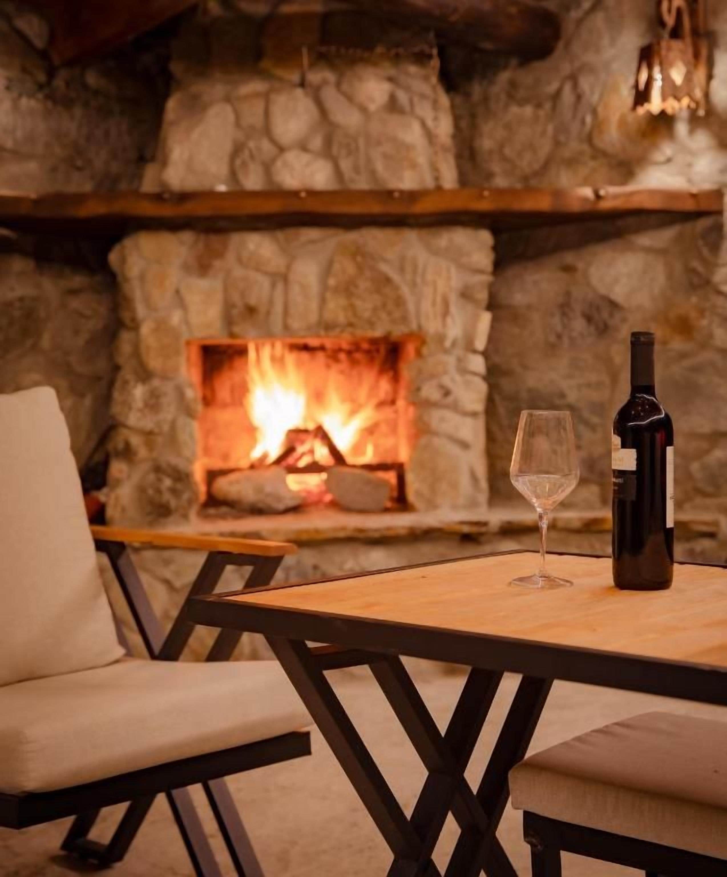 A stone fireplace with a fire, a bottle of wine, and a glass on a wooden table.