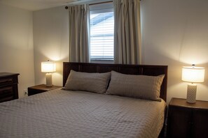 A neatly made bed with a quilted cover, flanked by two bedside tables with lamps, in a room with a window and curtains. - Waterfront Oasis - Stock Tank, Pool, Floating Dock & Kayaks (Hudson)