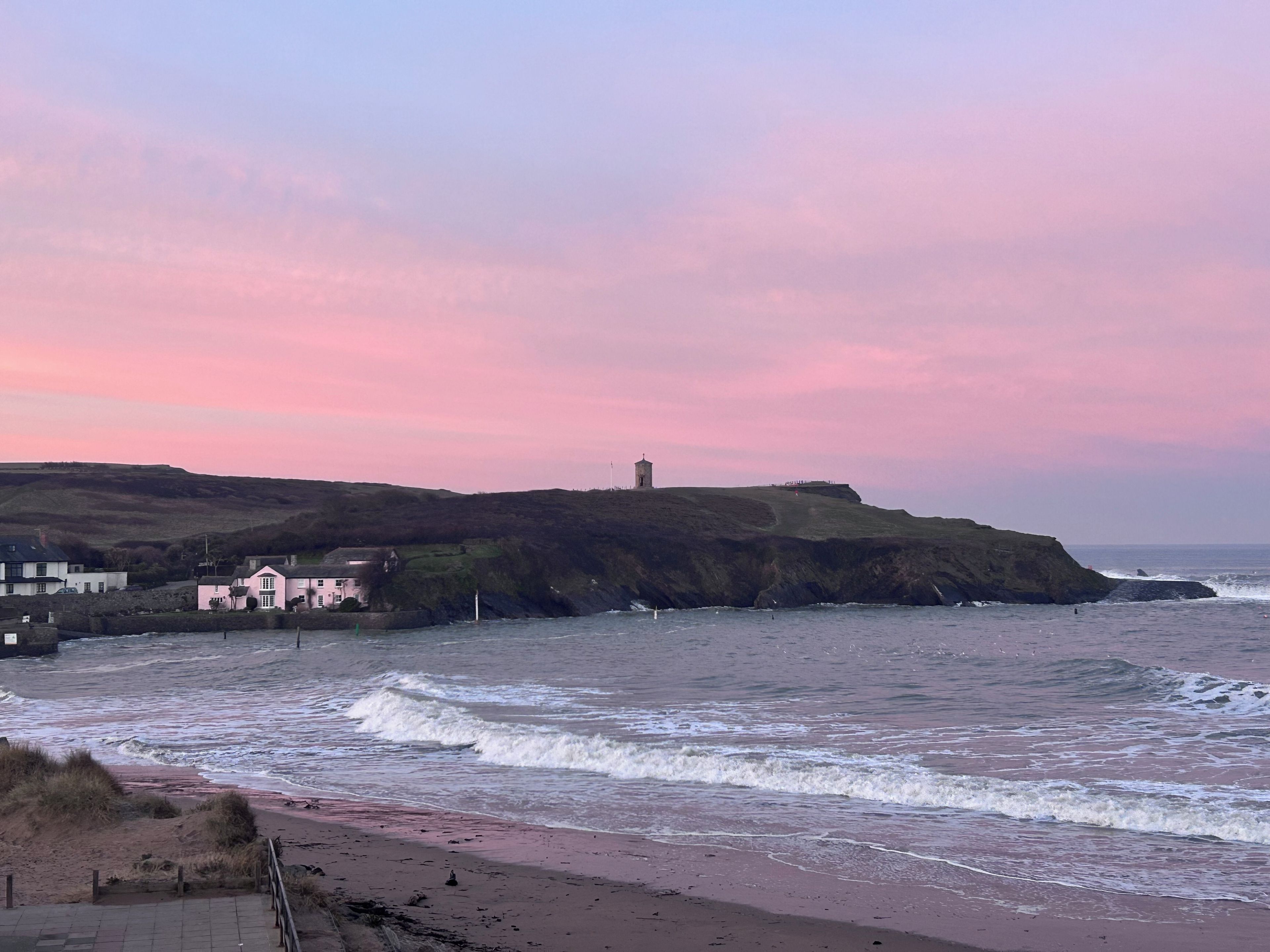 A coastal landscape with a lighthouse, buildings, and a beach.