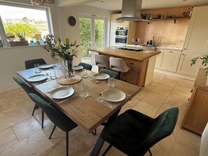 A modern kitchen with a central island, dining table set for six, and a view of the outdoors. - Family home in Bude 5 minutes from beach – garden, private parking & storage. (Bude)