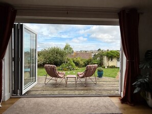 A room with a sliding glass door leading to an outdoor patio with two cushioned chairs and a small table. - Family home in Bude 5 minutes from beach – garden, private parking & storage. (Bude)