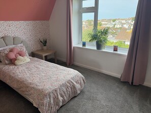 A bedroom with a bed, a small table, a potted plant, and a window with curtains. - Family home in Bude 5 minutes from beach – garden, private parking & storage. (Bude)