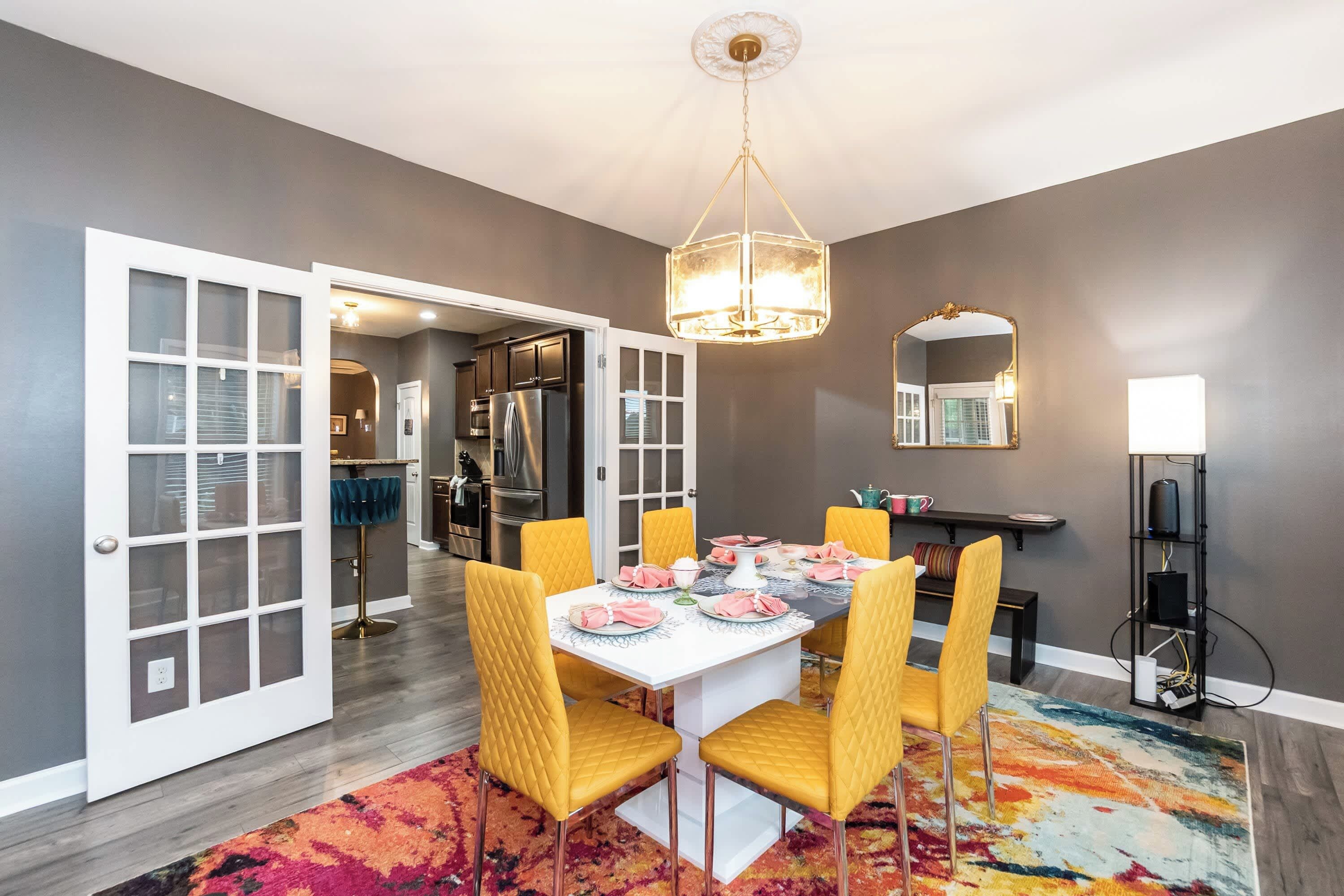 Un coin repas avec une table dressée pour six personnes, entourée de chaises jaunes, d’un tapis coloré et d’un lustre.