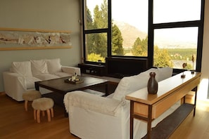 Un salon comprenant un canapé, une table basse et un pouf, un banc en bois et une grande fenêtre donnant sur les montagnes.