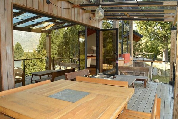 An outdoor wooden deck with tables and benches, a glass door leading to a room, and a view of trees and mountains.