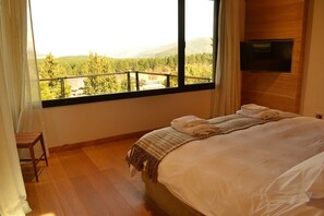 A bedroom with a large window, a bed with white linens, a wooden headboard, and a small wooden chair.