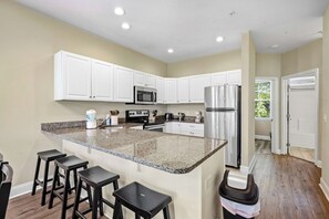 A modern kitchen with white cabinets, granite countertops, stainless steel appliances, and bar stools. - Salt Air & Sunshine – 3BR/3BA Myrtle Beach Townhouse, Steps to the Beach (Myrtle Beach)