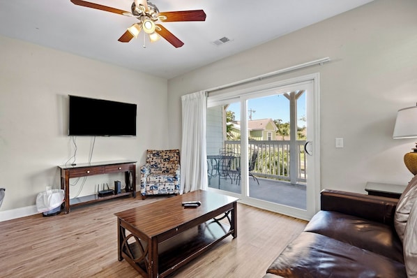 A living room with a wooden coffee table, a leather sofa, a TV mounted on the wall, and a sliding glass door leading to an outdoor area. - Salt Air & Sunshine – 3BR/3BA Myrtle Beach Townhouse, Steps to the Beach (Myrtle Beach)