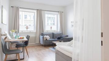 A modern bedroom with a bed, a sofa, a desk with chairs, and two windows with curtains.