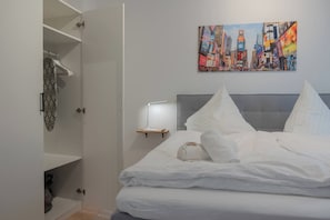 Ett hotellrum med en säng, en tavla som föreställer en stadsbild, ett nattduksbord med en lampa och en öppen garderob.