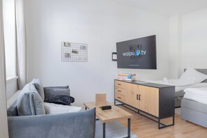 Ein modernes, helles Hotelzimmer mit Fernseher, Holzschrank, kleinem Tisch, einer Couch und einem Bett.