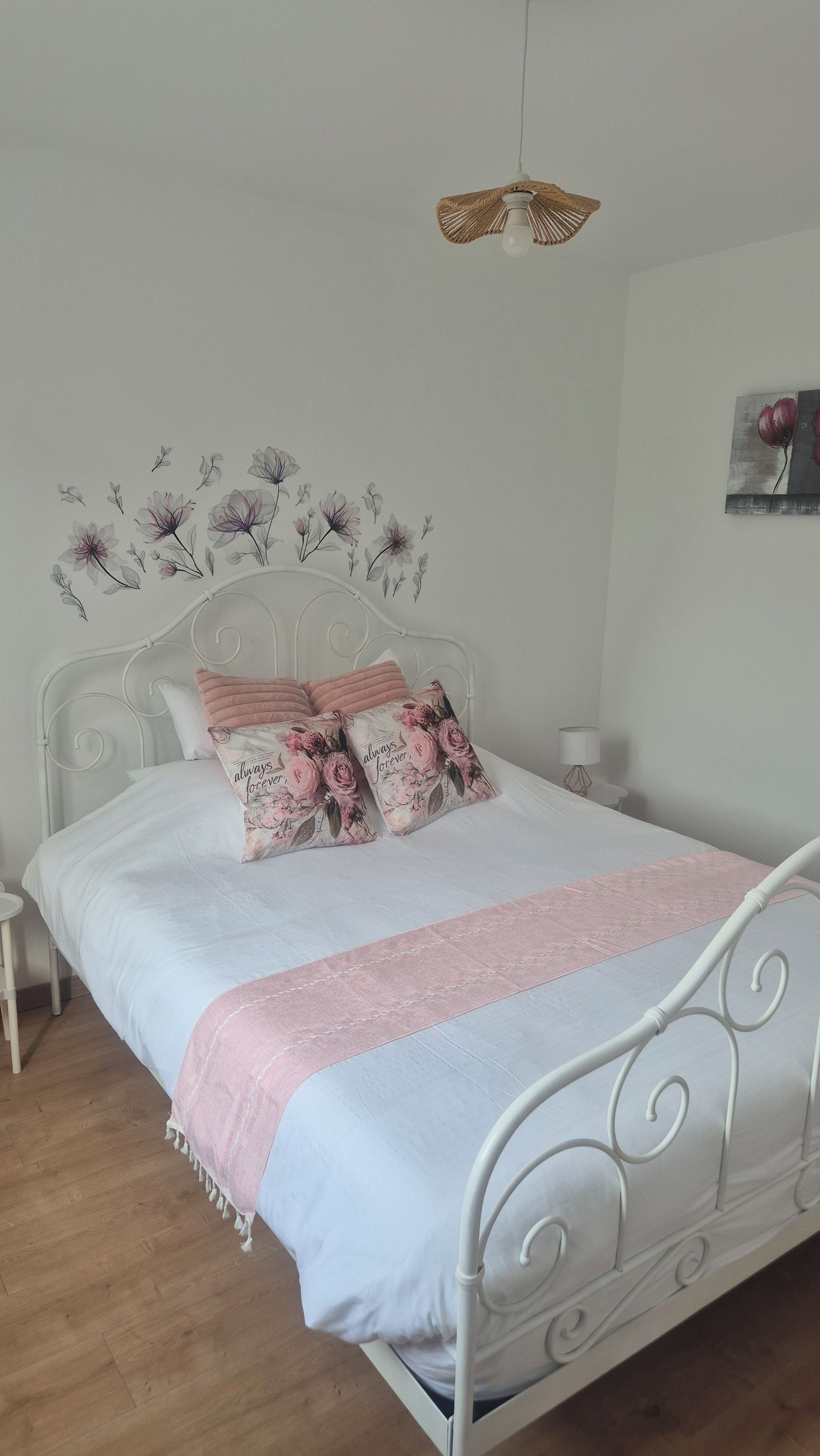 A neatly made bed with white bedding and pink accents, a white metal headboard, and a wall with floral wall art.