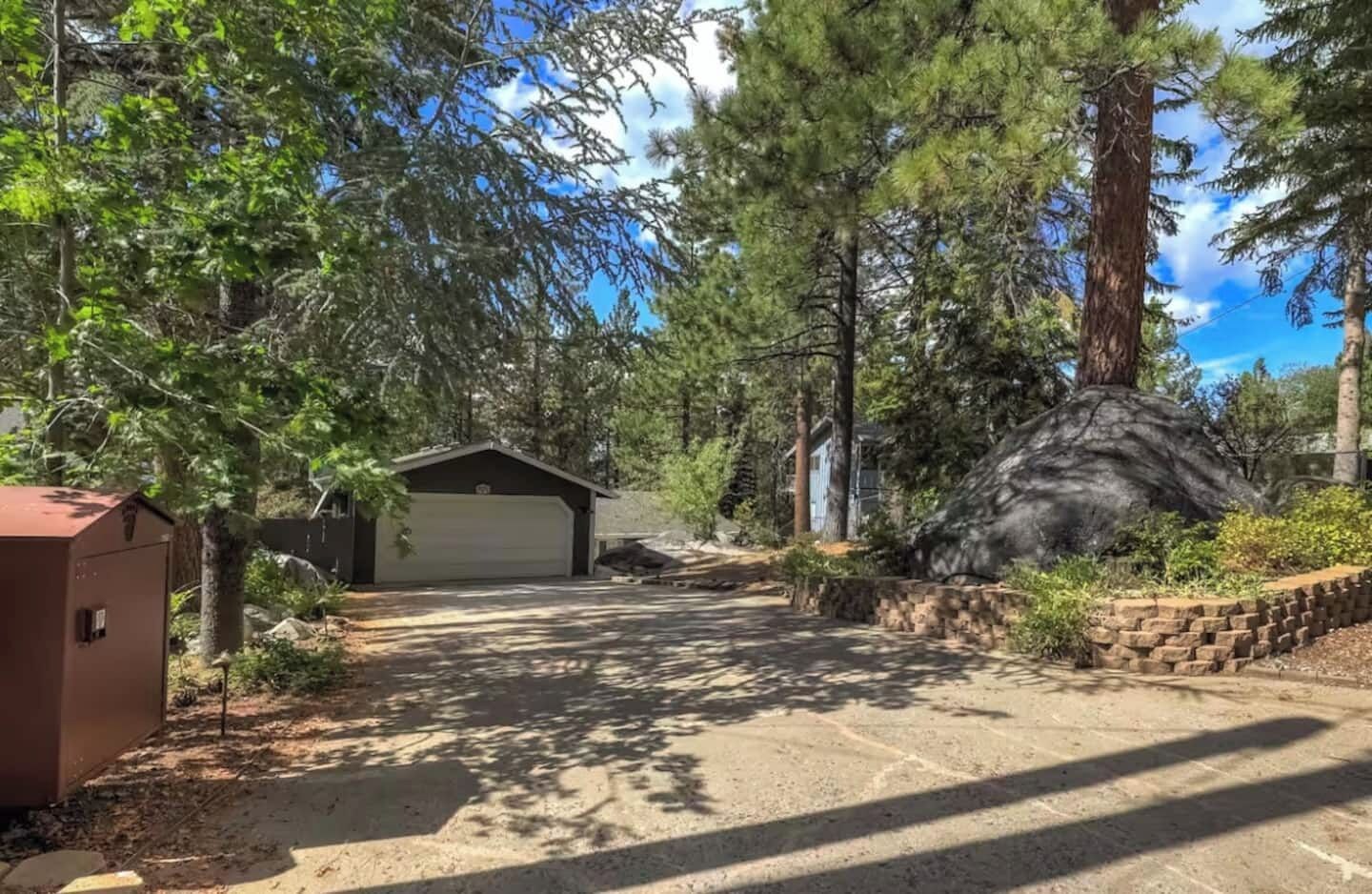 Une allée menant à une maison et aboutissant à un garage, entourée d’arbres et d’une grande formation rocheuse.