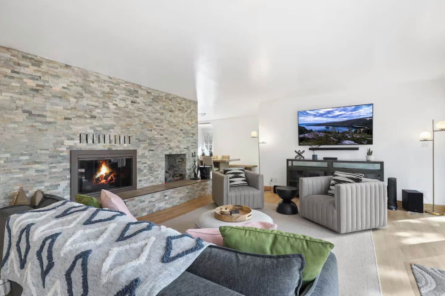 A modern living room with a stone fireplace, comfortable seating, a flat-screen TV, and a large window.