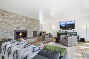 Ein modernes Wohnzimmer mit Kamin aus Stein, bequemen Sitzmöglichkeiten, einem Flachbildfernseher und einem großen Fenster.