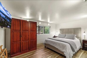 Downstairs bedroom with a spacious queen bed, wall mounted TV, large wardrobe, and soft lighting that creates a calm, comfortable place to unwind. There is a bathroom downstairs as well.