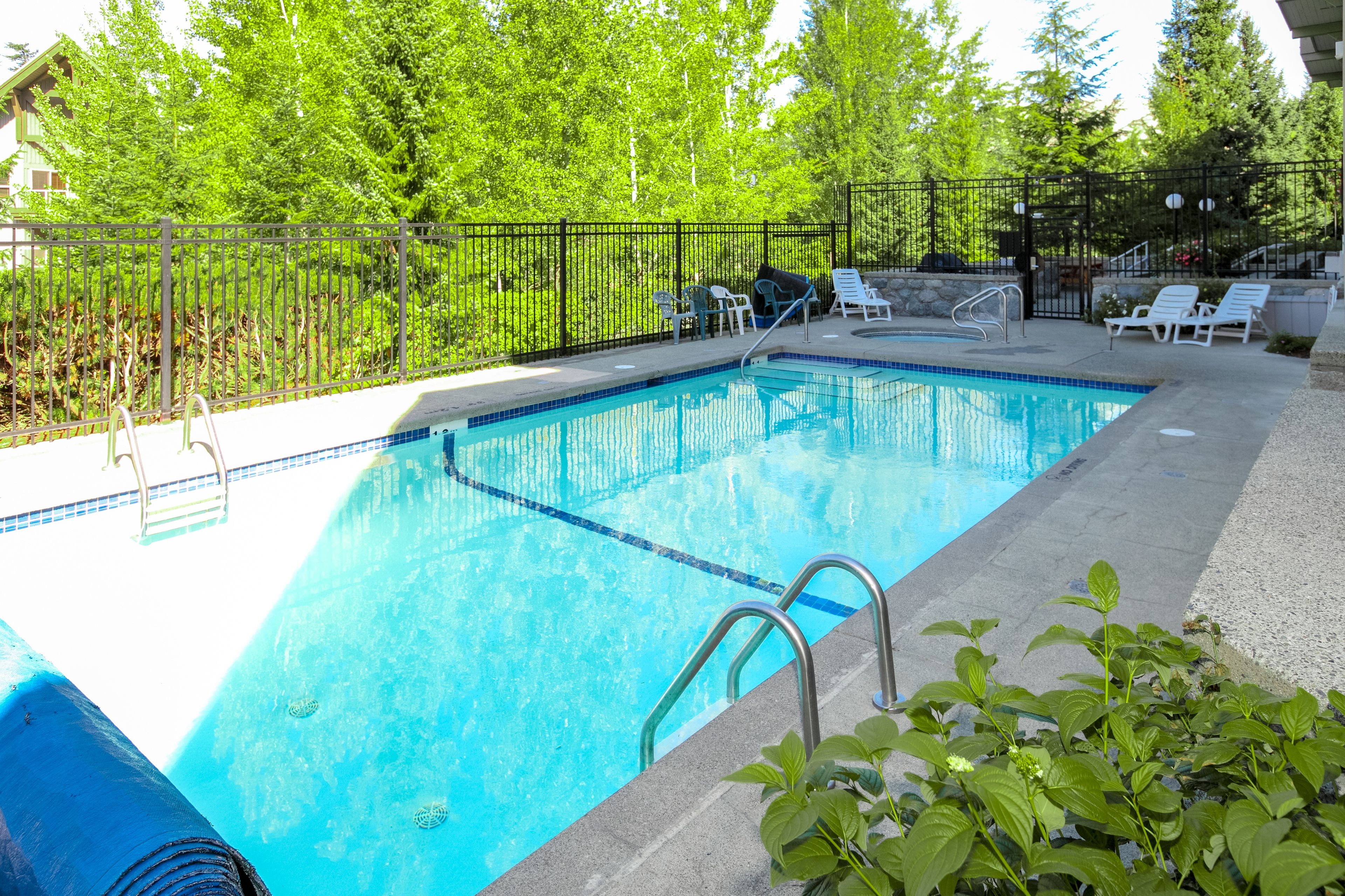 Ein rechteckiger Pool mit Fliesenrand, umgeben von einer niedrigen Steinmauer und einem Metallzaun.