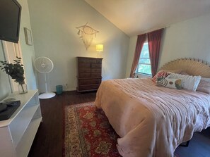 A bedroom with a bed, a dresser, a wall-mounted lamp, a fan, a window with curtains, and a decorative wall hanging. - Cozy 2-bedroom condo on St. Simons Island (St. Simons Island)