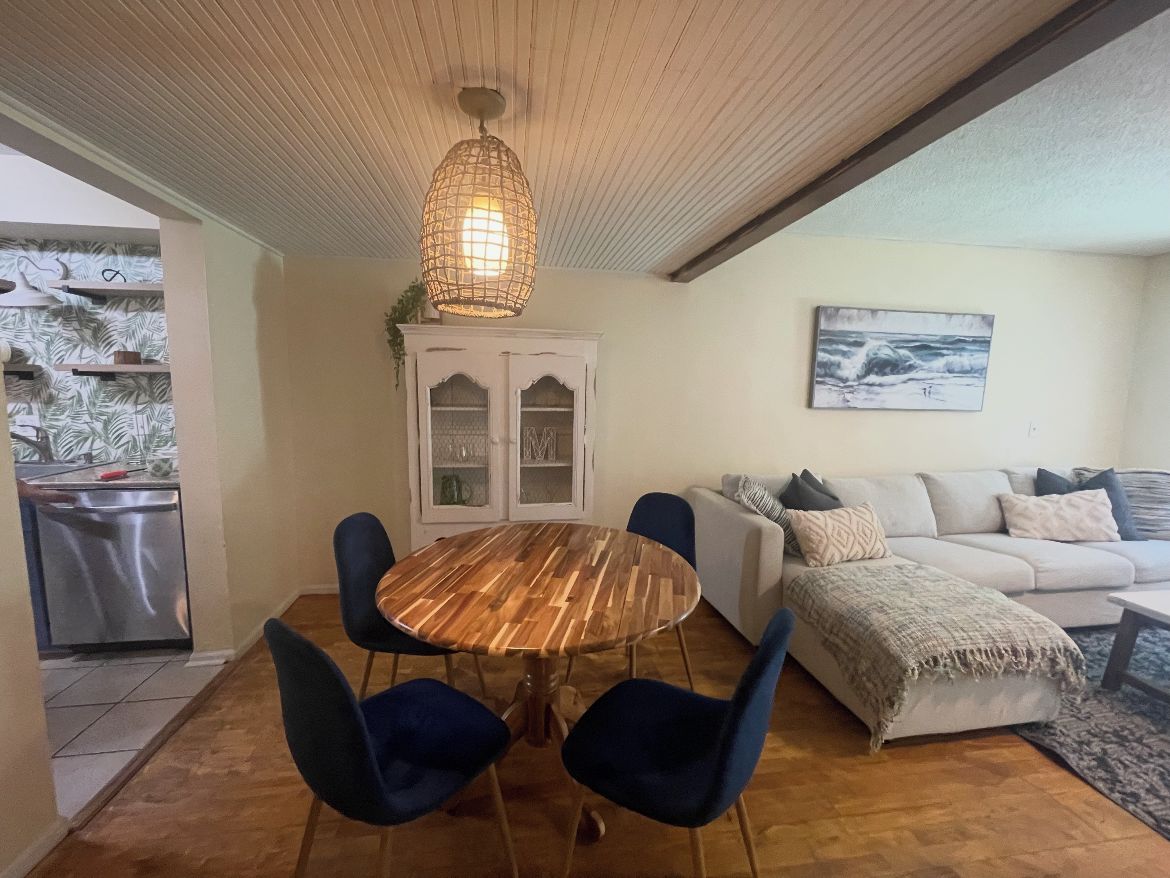 Ein Essbereich mit einem runden Holztisch, blauen Stühlen, einem weißen Schrank mit Glastüren und einem weißen Sofa mit Kissen.