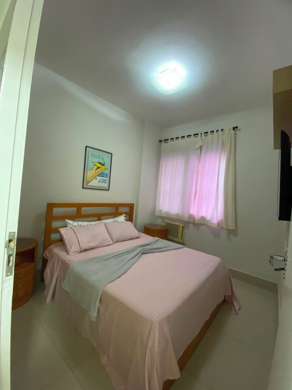 A bedroom with a bed, a bedside table, a wall-mounted TV, and a window with curtains.