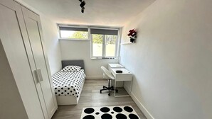 Een kleine, smalle kamer met een bed, een bureau en een stoel. Er is een raam dat natuurlijk licht biedt.