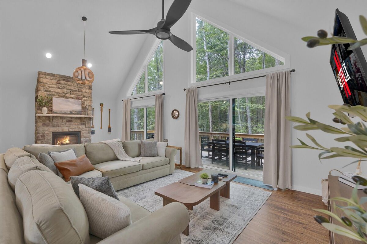 A spacious living room with a fireplace, a ceiling fan, and a large window overlooking a deck.